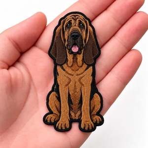 May include: An embroidered patch featuring a tan and brown Bloodhound dog sitting in a forward-facing position. The dog has long, dark brown ears, a black muzzle, and a pink tongue visible. The patch has a thick black border outline.
