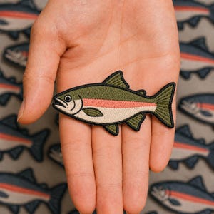 May include: An embroidered fish patch featuring an olive green back, a cream-colored belly, and a horizontal pink stripe along the side. The fish has a black stitched outline, a small white eye with a black pupil, and detailed fin textures.