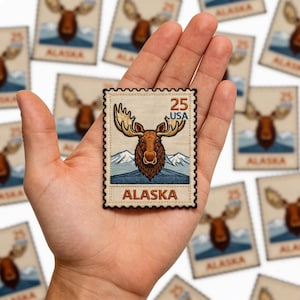May include: A rectangular patch featuring a moose with large antlers, mountains, and the word "ALASKA". The patch has a postage stamp design with the text "25 USA". The patch is held in a hand.