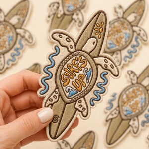May include: An embroidered patch featuring a sea turtle on a surfboard with the text SURF'S UP in yellow. The design includes light green, cream, and blue wave accents, with a small yellow sun icon at the top, all outlined in dark brown thread.