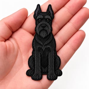 May include: An embroidered patch featuring a black Schnauzer dog sitting in an upright position. The design showcases detailed black and dark gray thread work, highlighting the dog's characteristic beard, pointed ears, and textured fur coat.