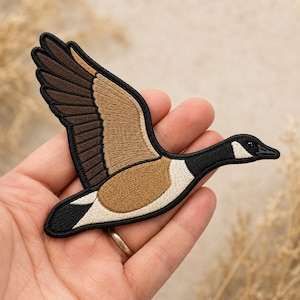 May include: An embroidered patch shaped like a Canada goose in flight. The goose features a black head and neck with a white cheek patch, brown and tan wings, and a white underbelly. The entire design is outlined with a thick black border.