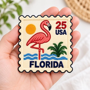 May include: A square embroidered patch with scalloped edges featuring a pink flamingo standing in blue water. The design includes a yellow sun, a green palm tree, and the text 25 USA and FLORIDA in dark blue thread on a cream background.