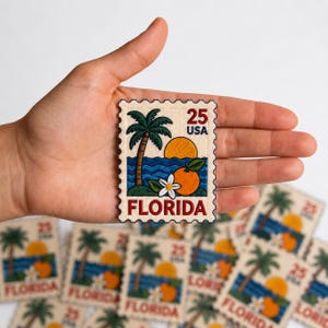 May include: A rectangular embroidered patch shaped like a postage stamp featuring a palm tree, a yellow sun, blue ocean waves, an orange fruit, and a white blossom. The text 25 USA and FLORIDA is stitched in red and dark blue on a cream background.