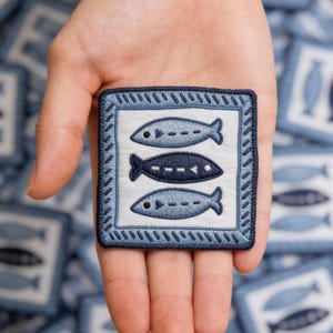 May include: A square, embroidered patch featuring three stylized fish in shades of blue. The fish are on a white background, framed by a blue border with a decorative stitch. The patch is held in a hand.