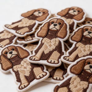 May include: Embroidered patches featuring a Cavalier King Charles Spaniel dog design. The patches have a white border, with the dog's fur in shades of brown and tan. The dog is sitting, with its ears flopped down.