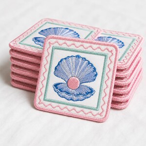 May include: A stack of square embroidered patches featuring a white background with a central blue seashell containing a pink pearl. Each patch has a light green square border and a scalloped pink stitched edge, creating a decorative finish.
