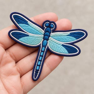 May include: An embroidered dragonfly patch featuring a dark blue border and body with wings in shades of light blue, turquoise, and dark blue. The insect design has a segmented body and detailed wing patterns outlined in white thread.