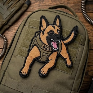 May include: An olive green tactical backpack featuring a large embroidered patch of a German Shepherd dog wearing a K9 harness. The dog is depicted with its mouth open, showing teeth and a pink tongue, set against the textured fabric of the bag.