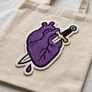 May include: A beige canvas tote bag featuring a large embroidered patch of a purple anatomical heart pierced by a silver sword with a gold hilt. The embroidery includes black outlines and a small purple teardrop shape below the heart.
