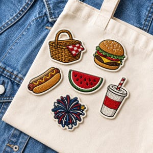 May include: A cream-colored canvas tote bag featuring six embroidered patches: a brown picnic basket with a red and white checkered cloth, a cheeseburger, a hot dog with yellow mustard, a watermelon slice, a drink cup, and a blue firework.