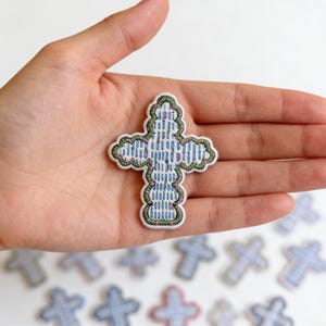May include: Embroidered cross patch with a blue and white checkered pattern, outlined in green and pink. The cross is held in a hand, with several similar patches visible in the background. The patch is a religious symbol.