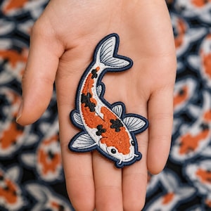 May include: An embroidered iron-on patch shaped like a koi fish. The fish features a white body with vibrant orange and black patches, detailed with white fins and a dark navy blue border. The design captures the fish in a curved swimming motion.