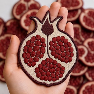 May include: An embroidered patch shaped like a halved pomegranate, featuring deep red seeds and a cream-colored rind with a dark maroon border. The design displays three distinct clusters of seeds and a stylized stem at the top of the fruit.