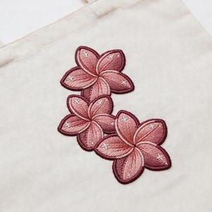 May include: Cream-colored tote bag featuring an embroidered floral design. The design showcases three pink flowers with dark red outlines and white star-shaped accents. The flowers are arranged in a cascading pattern.