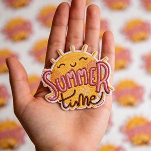 May include: An embroidered patch featuring a yellow sun with a smiling face and pink cheeks. The text "SUMMER time" is stitched in pink and dark brown thread across the center of the sun, which has yellow rays extending outward.