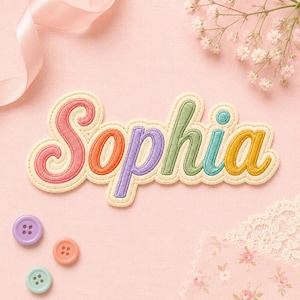 May include: An embroidered fabric patch featuring the name Sophia in a cursive font. Each letter is a different color: pink, orange, purple, green, teal, and yellow. The patch has a cream-colored border and is surrounded by three small buttons.
