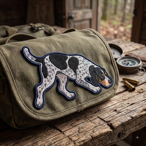 May include: An olive green canvas messenger bag featuring a large embroidered patch of a white, black, and tan hunting dog in a tracking pose. The dog has a spotted coat, dark ears, and a tan muzzle, outlined in a dark blue stitched border.