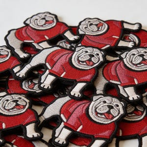 May include: Embroidered patches featuring a bulldog wearing a red sweater. The patches have a white and gray color scheme, with the bulldog's face and paws in white. The red sweater has a zipper detail. The patches are scattered on a white surface.
