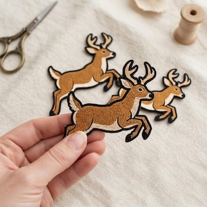 May include: Three embroidered iron-on patches featuring a leaping deer with brown fur, a cream-colored underbelly, and light tan antlers. Each patch is outlined in a thick black border, showcasing detailed stitching that defines the animal's form.