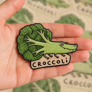May include: Embroidered patch featuring a green broccoli-shaped alligator with the word "CROCCOLI" on a tan background. The patch has a black border and is held in a hand.
