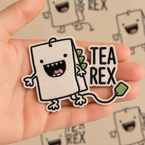 May include: Embroidered patch featuring a cartoon tea bag character with a dinosaur tail and the text "TEA REX". The patch is off-white with black outlines, a green tail, and a pink mouth. The design is whimsical and playful.