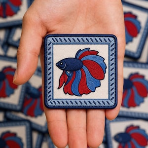 May include: A square embroidered patch featuring a betta fish with blue and red fins on a cream-colored background. The patch is framed by a dark blue border with a decorative stitched pattern, creating a distinct contrast against the center.