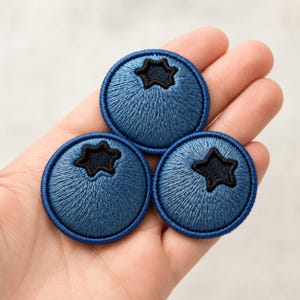 May include: Three circular embroidered patches shaped like blueberries. Each patch features a vibrant blue thread texture radiating from a dark navy blue star-shaped center, finished with a solid dark blue stitched border around the circumference.