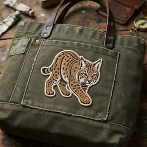 May include: An olive green canvas tote bag featuring a large embroidered patch of a brown and black spotted lynx on the front pocket. The bag has dark brown leather handles secured with metal rivets and a frayed edge on the front pocket fabric.