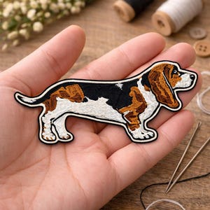 May include: Embroidered patch featuring a Basset Hound dog. The patch has a white border and depicts the dog in brown, black, and white. The dog is standing with its long ears hanging down. The patch is likely for clothing or accessories.