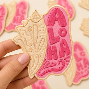 May include: An embroidered patch featuring a beige seashell on the left side and the word ALOHA in bold pink lettering on the right side. The design includes decorative swirls and dots, with a light pink background behind the text.