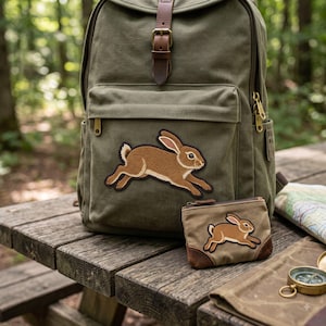 May include: An olive green canvas backpack and a matching small zippered pouch, both featuring an embroidered brown rabbit patch. The backpack has a brown leather strap closure, and the pouch has brown leather trim at the bottom corners.