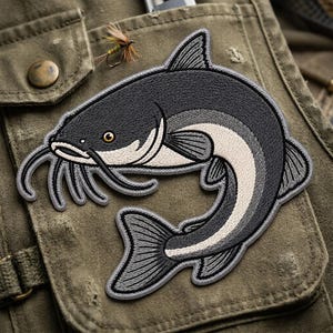 May include: An embroidered patch featuring a dark gray and cream-colored catfish with long whiskers, a prominent dorsal fin, and a curved tail. The fish is depicted in a swimming motion with detailed stitching throughout its body and fins.
