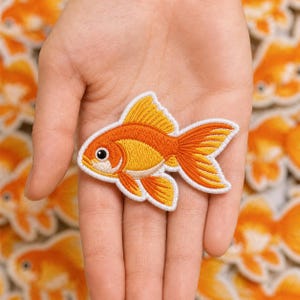 May include: An embroidered patch shaped like a goldfish with vibrant orange and yellow thread, featuring a white border, a black eye, and detailed fin stitching. The design captures the classic form of a goldfish with a rounded body and fins.