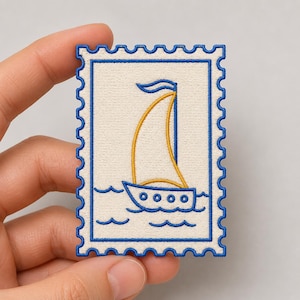 May include: A rectangular embroidered patch shaped like a postage stamp with a cream-colored background. The design features a blue-outlined sailboat with a yellow sail and blue waves at the bottom, all bordered by a scalloped blue edge.