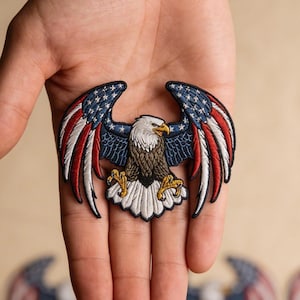 May include: Embroidered patch featuring a bald eagle with outstretched wings. The wings display the American flag design with red and white stripes and blue stars. The eagle has a white head, brown body, and yellow talons.
