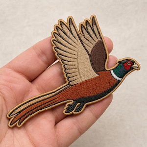 May include: An embroidered patch featuring a pheasant in flight. The bird has a red face, a dark green neck with a white ring, a brown and tan body with detailed wing feathers, and a long, pointed tail. The entire patch has a gold-colored border.