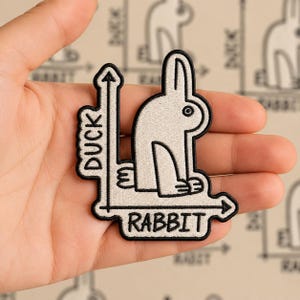 May include: An embroidered patch featuring a white rabbit design with the text "DUCK" and "RABBIT" in black. The patch has a black border and is held in a hand.
