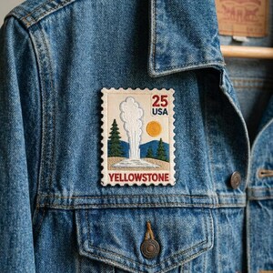 May include: A rectangular embroidered patch featuring a Yellowstone geyser design attached to a blue denim jacket. The patch displays a white geyser plume, green pine trees, a yellow sun, and the text 25 USA YELLOWSTONE in red and blue.