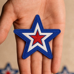 May include: An embroidered star-shaped patch featuring a layered design with a solid red center star, a white middle star border, and a thick blue outer star border. The embroidery has a textured finish with visible stitching throughout the design.