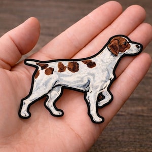May include: Embroidered patch featuring a white dog with brown spots and a brown head. The dog is outlined in black. The patch is likely for clothing or accessories.