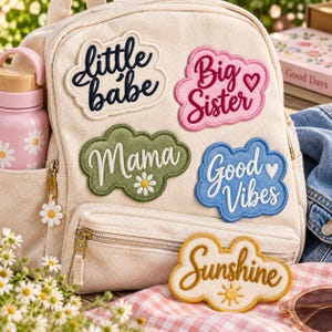 May include: A cream-colored canvas backpack decorated with five cloud-shaped embroidered patches featuring the text little babe, Big Sister, Mama, Good Vibes, and Sunshine, along with a pink water bottle with white daisy patterns and a daisy zipper pull.