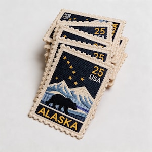 May include: A stack of embroidered patches resembling vintage postage stamps. Each patch features a black bear silhouette, mountains, and the word "ALASKA" in gold lettering. The patches also display the text "25 USA" and a constellation of stars.