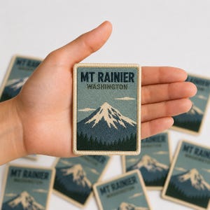 May include: A rectangular embroidered patch featuring a landscape of Mount Rainier in Washington. The design displays a snow-capped mountain peak in white and blue, a dark green forest at the base, and a light blue sky with clouds. The text reads MT RAINIER WASHINGTON.