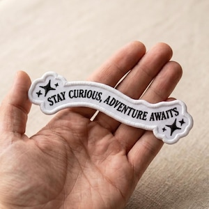 May include: A white embroidered patch with the text "STAY CURIOUS, ADVENTURE AWAITS" in black. The patch has a cloud-like shape with black star designs at each end. The patch is held in a hand.