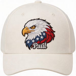 May include: Cream-colored baseball cap featuring an embroidered eagle head design. The eagle has white feathers, a yellow beak, and a patriotic red, white, and blue color scheme. The name "Paul" is embroidered below the eagle.