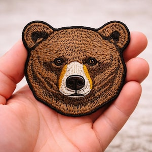 May include: Embroidered patch featuring a detailed bear face. The bear has brown fur with black outlining, a white muzzle, and yellow accents. The patch is designed to be sewn or ironed onto fabric, suitable for jackets, bags, or other items.