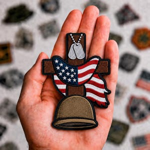 May include: An embroidered patch featuring a brown cross, a tan military helmet at the base, a draped American flag, and a pair of silver-colored dog tags hanging from the top of the cross, all outlined in black thread on a fabric backing.