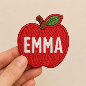May include: Embroidered red apple patch with the name "EMMA" in white block letters. The apple has a brown stem and a green leaf. This patch is likely for clothing or accessories.