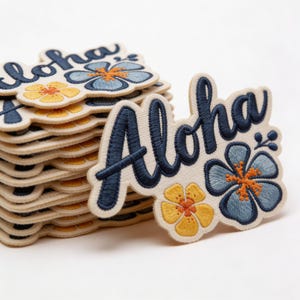 May include: Embroidered patches with the word "Aloha" in navy blue script, accompanied by blue and yellow hibiscus flower designs. The patches have a cream-colored background and are stacked, showcasing the design.